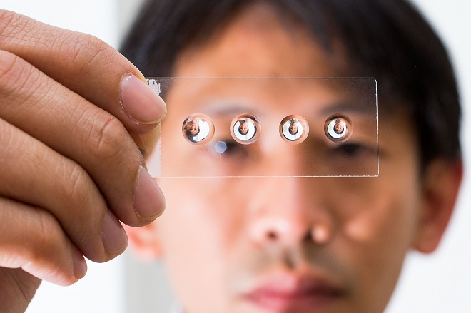 A set of droplet lenses on a microscope coverslip held up by ANU researcher Steve Lee